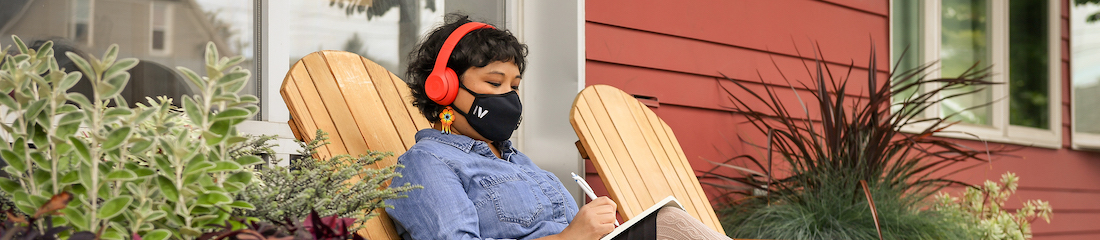 A Filipinx woman with a filtering face mask sits outside a cafe and writes in a notebook. She is wearing headphones and in front of a window, with potted plants all around. Mask optional.