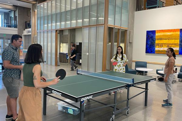 employees playing ping pong in the atrium of the center building with the library in the background