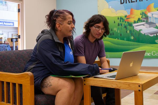 two student workers, a young man with long hair and classes, and a woman in her thirties with pink hair and an LCC shirt, work in the admissions office