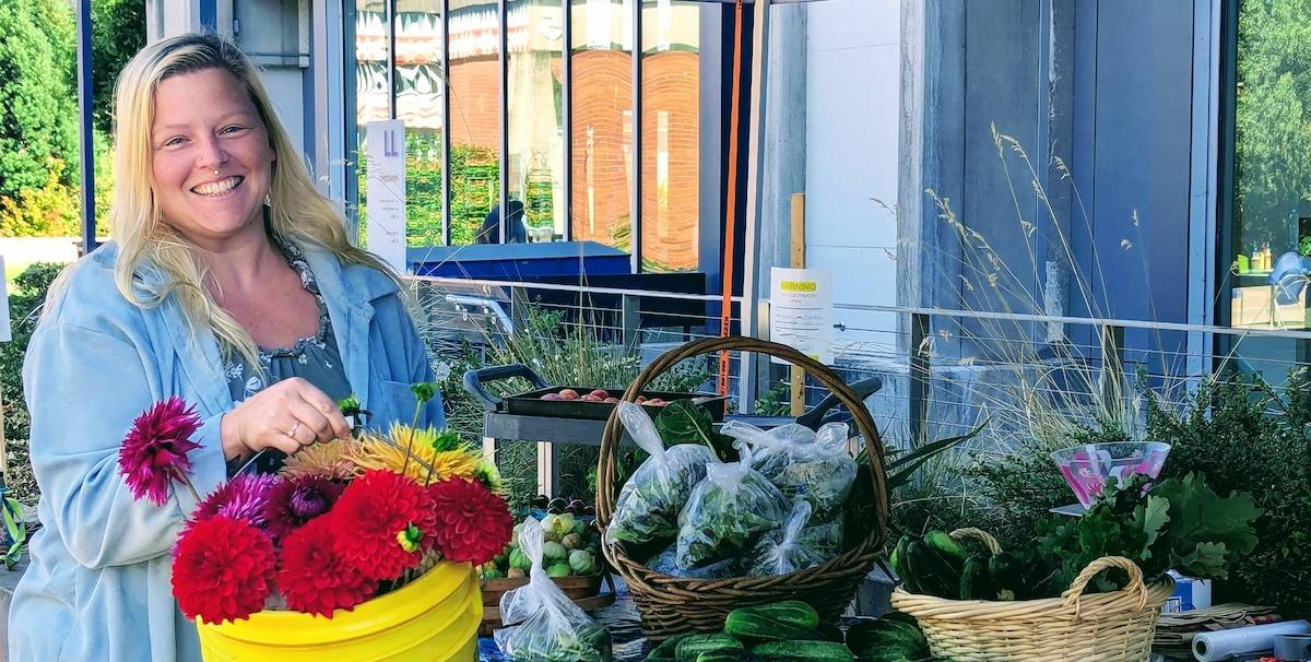 a Lane employee, a smiling blonde woman with a nose piercing, holds a basket of flowers and smiles at a pop up produce market
