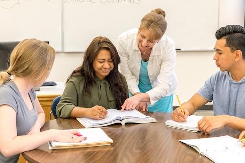 a business instructor smiles as she leans over a table to point out something in a textbook to students during class