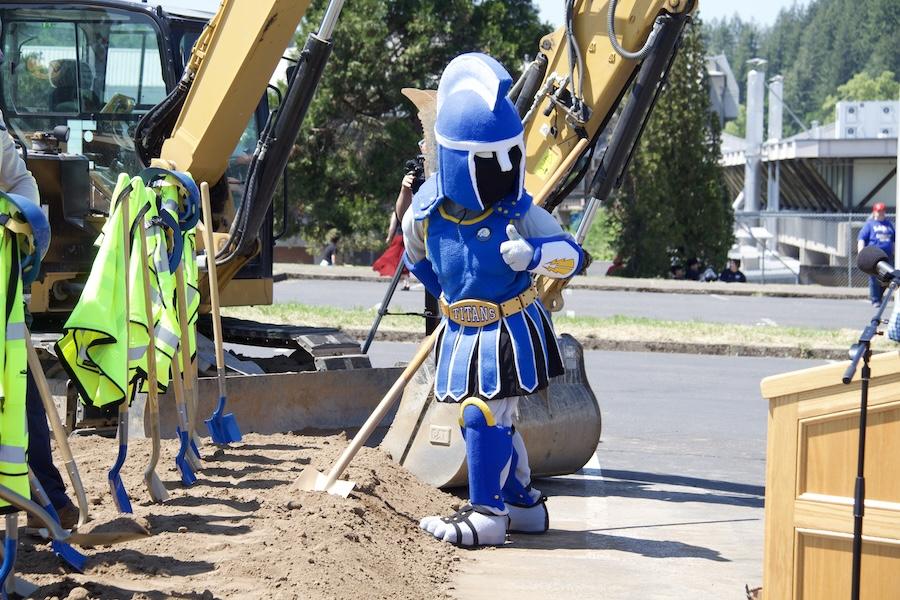 Ty the Titan mascot giving a thumbs up to the camera, while posing with a shovel at a building groundbreaking near construction machinery