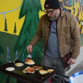 an art instructor cooks pancakes for an event next to a large colorful mural on campus