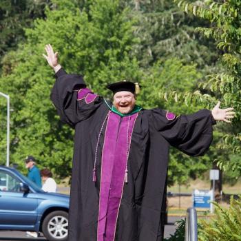 a manager, a smiling man in graduation robes, smiles with hands outstretched in a goofy pose at Commencement