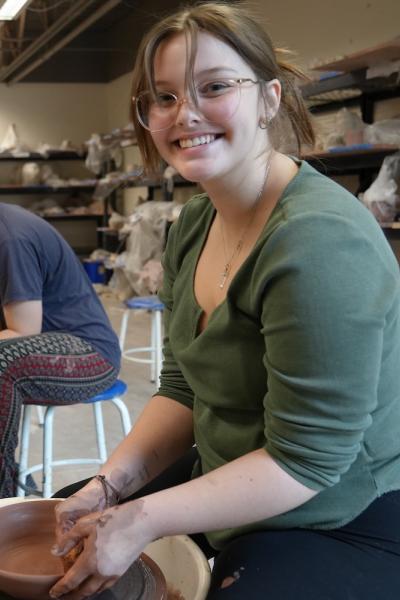 A young woman looks up from a pottery wheel she's working at smiling